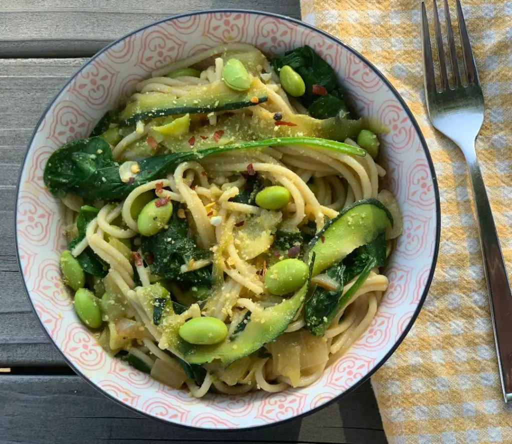 Gorgeous bowl of pasta with edamame, spinach, and zucchini ribbons, topped with vegan parmesan, with a yellow plaid napkin and fork adjacent, shot on a picnic table.
