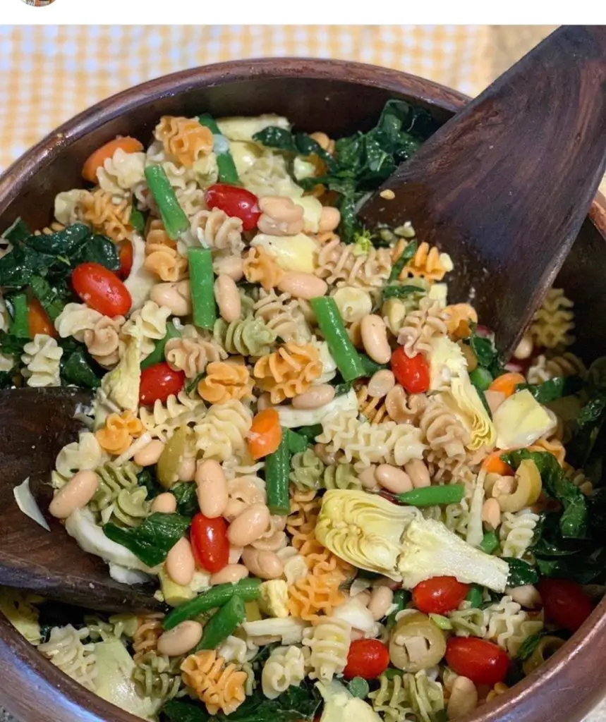 Gorgeous colorful pasta salad with artichoke hearts, tomatoes, olives, green beans, and white beans in a tricolor pasta mix. Shown in brown wooden salad bowl with brown wood salad tongs.
