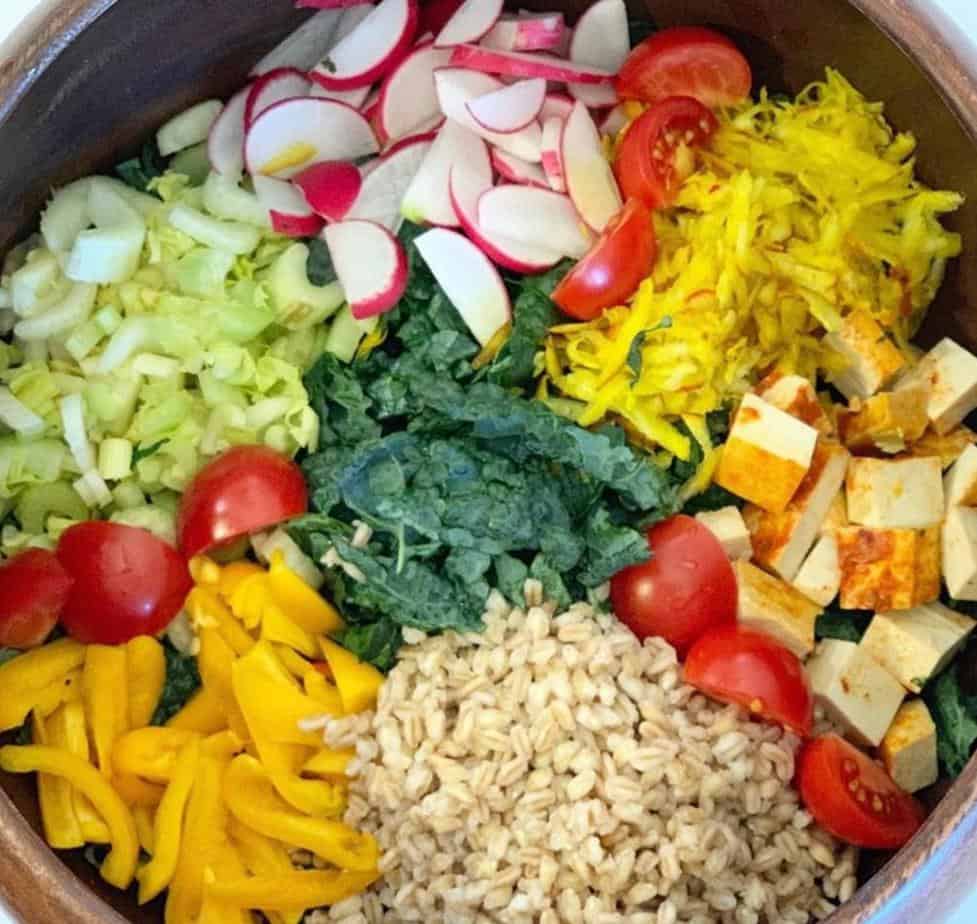 Rainbow salad with all ingredients neatly placed in salad bowl in clockwise placement:radishes, beets, tofu, tomatoes, farro, peppers, tomatoes, celery, and kale in the middle. No dressing shown.