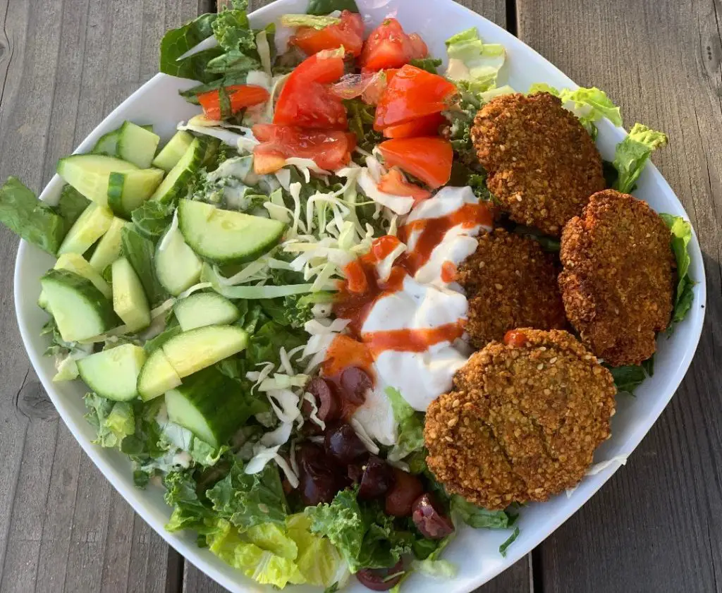 Falafel salad with cucumbers, tomatoes, and tzatziki in white bowl on picnic table.