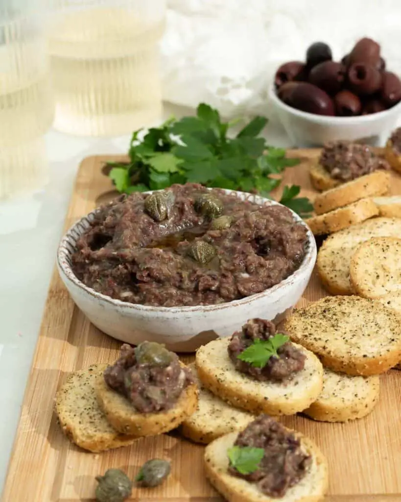Eggplant tapenade in bowl with herbed crackers and parsley on cutting board around the bowl.