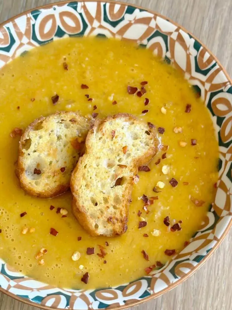 Close up of carrot and lentil soup in patterned bowl, topped with large croutons and red pepper flakes.