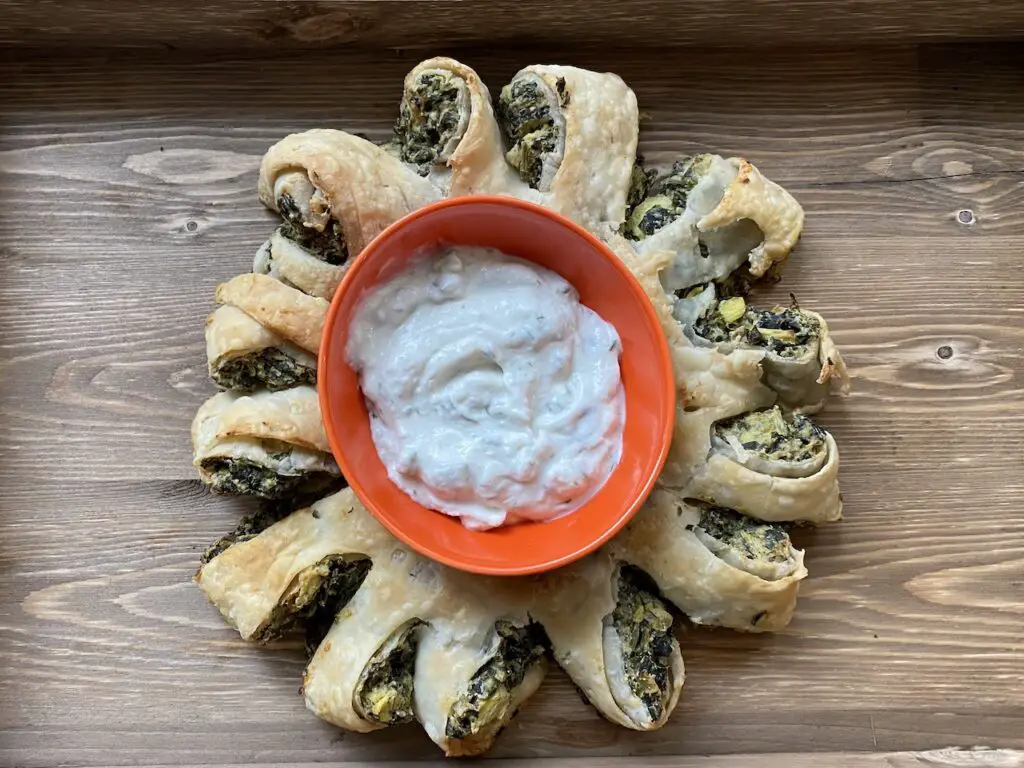 Puff pastry wreath on wood cutting board with orange bowl full of vegan tzatziki in the middle of the wreath.