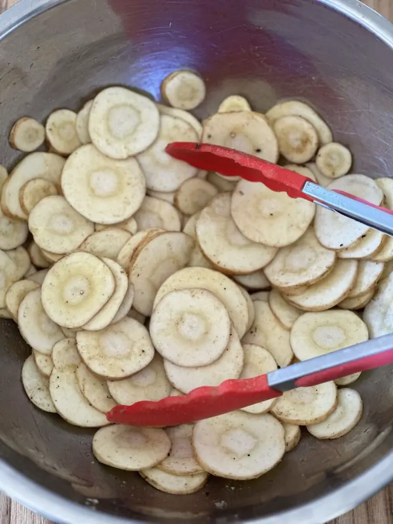 Tongs are mixing oil and spices with raw parsnip chips in bowl.