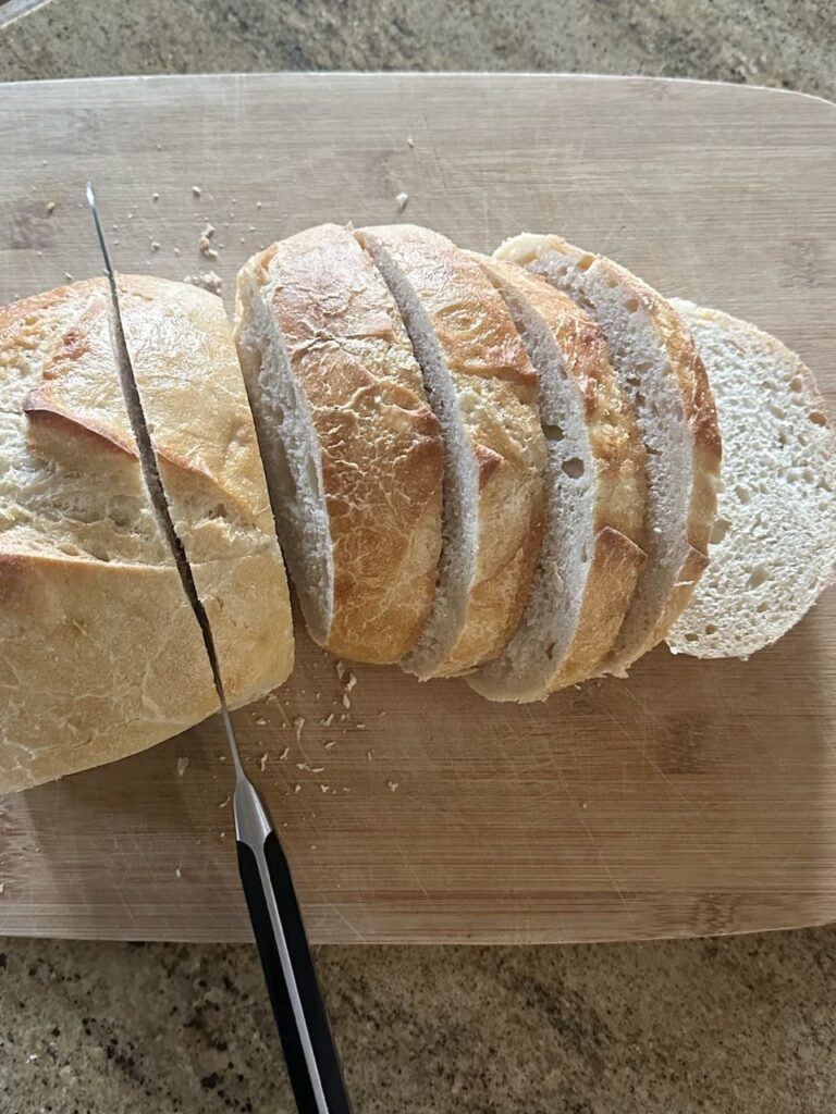 Loaf of thick french bread being cut into 1 inch thick slices on cutting board.