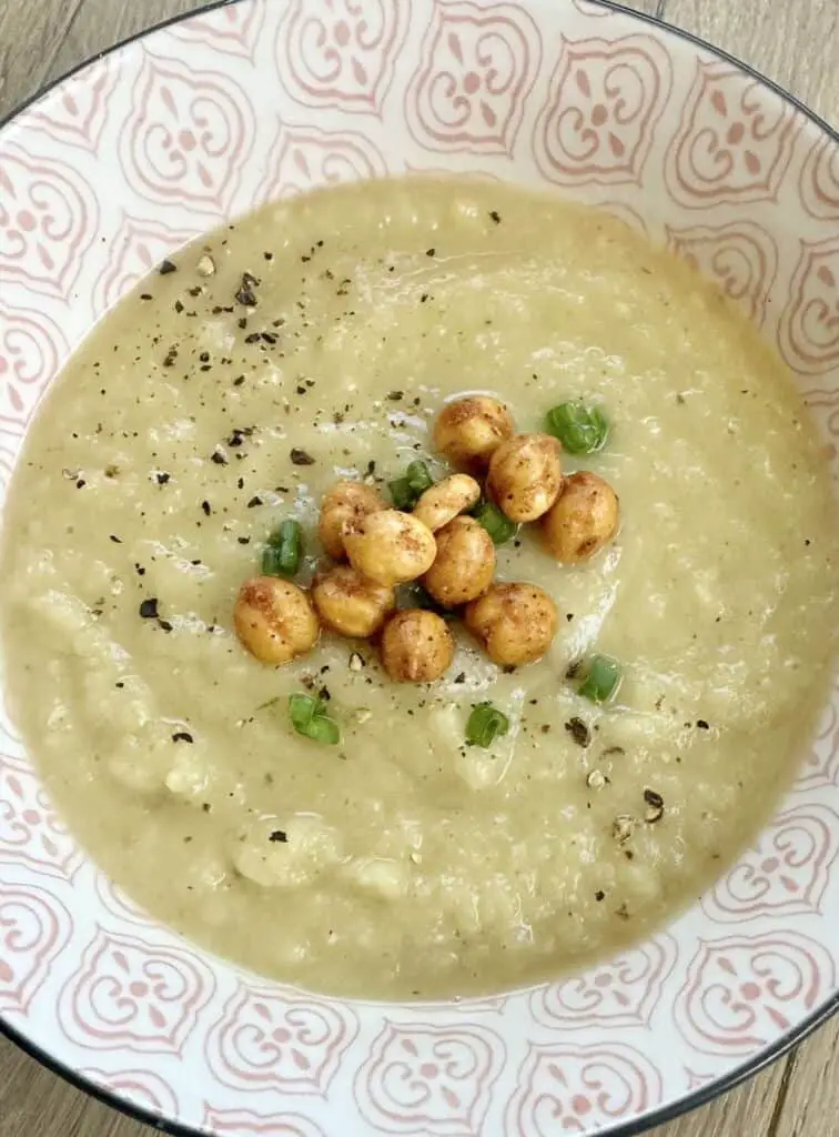 Vegan Celery Root Soup shown in pink and white printed bowl and topped with black pepper, roasted chickpeas, and green onions.