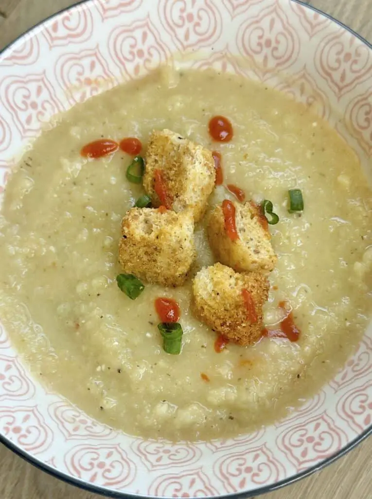 Celeriac soup in pink printed bowl topped with croutons and sriracha and green onions.