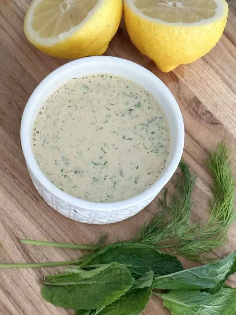Lemon Herb Tahini Dressing on cuting board with lemon, mint, dill, and parsley.