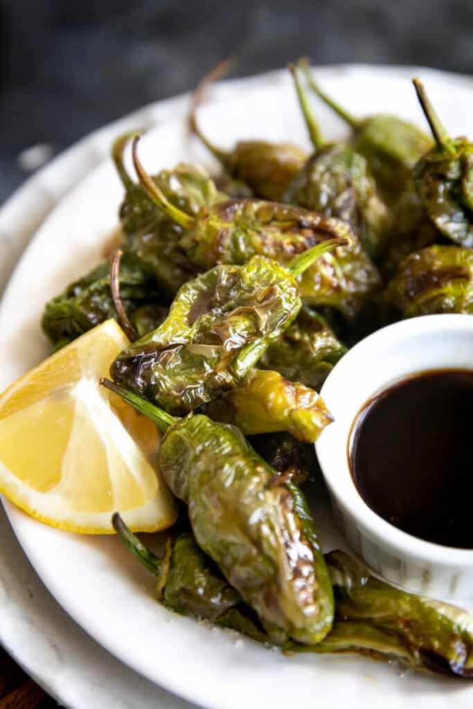 Close up of shishito peppers, on a plate with lemon wedges and a dipping sauce.