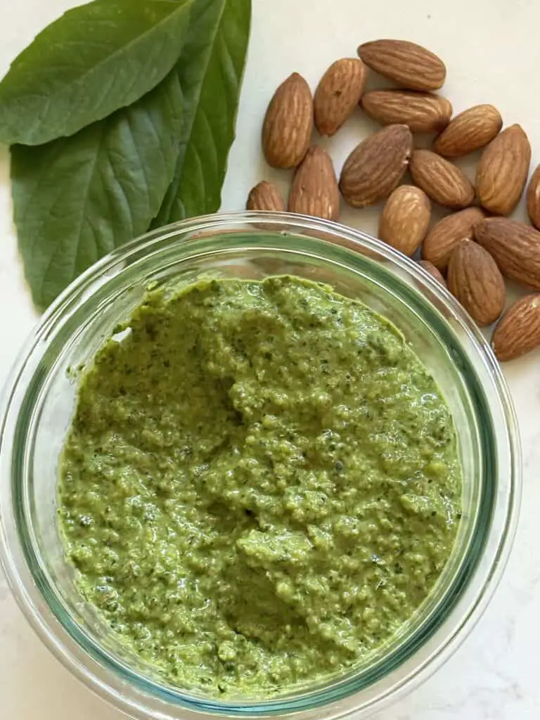 Vegan almond pesto in jar with toasted almonds and basil adjacent.