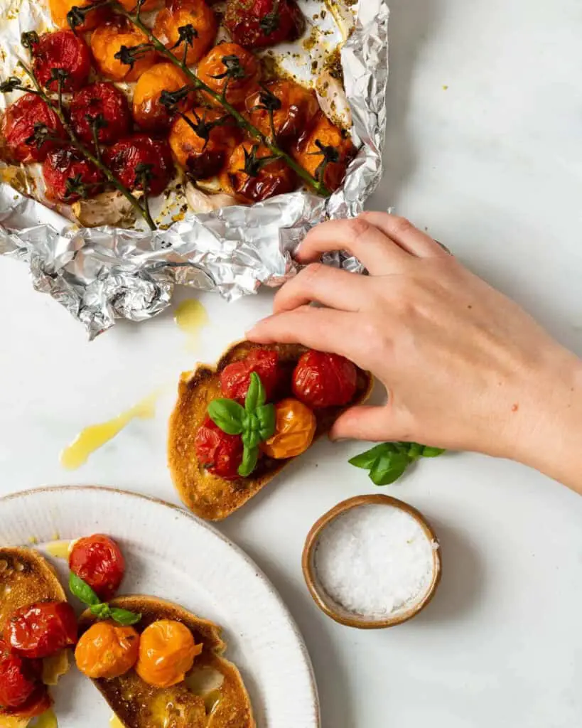 Air fryer tomatoes, some in foil, some no a plate over toast, and a hand grabbing one of the toasts on the counter.