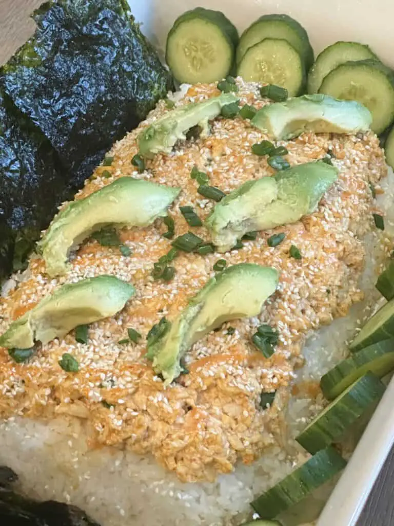 Vegan sushi bake in pan topped with avocado and shown with cucumbers on the right side of pan and seaweed sheets on left side.