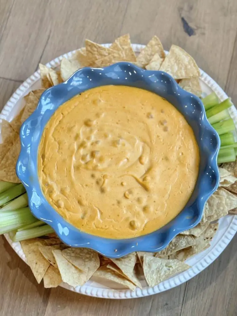 Vegan buffalo chickpea dip in light blow dish, served with tortilla chips and celery sticks. Photograph on wood floor.