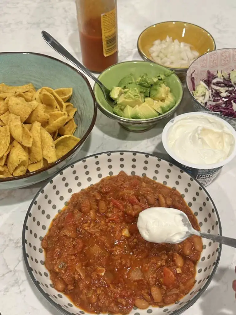 Chili bar: bowl of chili and a variety of topping bowls, including corn chips, vegan sour cream, avocado, cabbage,  onions, and hot sauce.